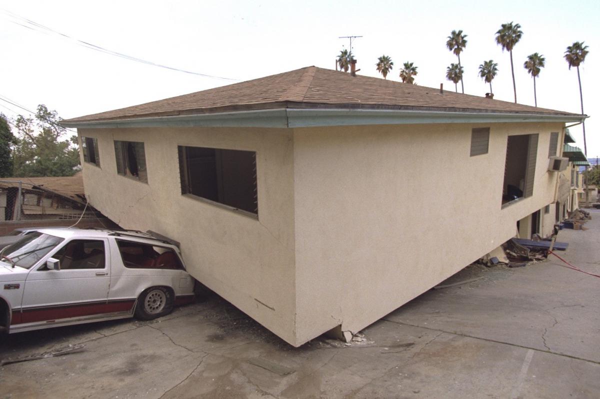 A one-story structure leaning on the ground partially crushing a car underneath with palm trees in the background.