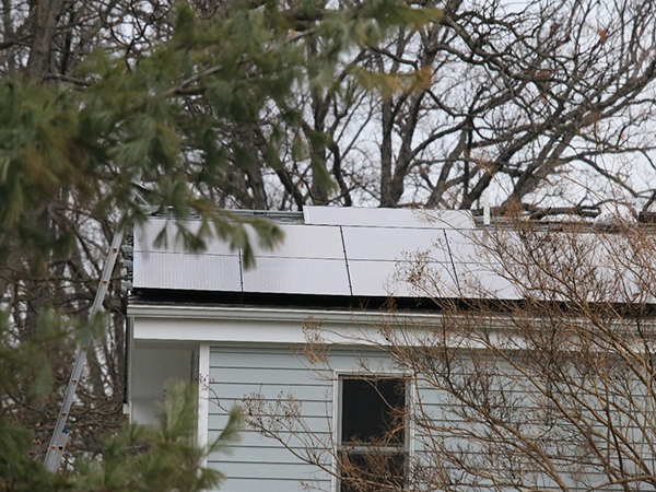 Solar panels have been installed on the roof of a home in a wooded area. A ladder on the left side of the home provides access to the roof for the workers installing the solar panels.