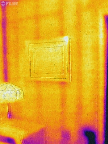 Thermal image of a wall, in which the studs and exterior outlet are visible, as is heat loss in the corner. The table lamp and picture frame on the wall are also visible.