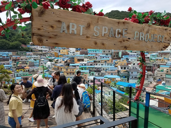 People stand on a wooden deck enclosed by a metal railing overlooking homes. A sign stating, “Art Space Project,” is visible in the foreground.
