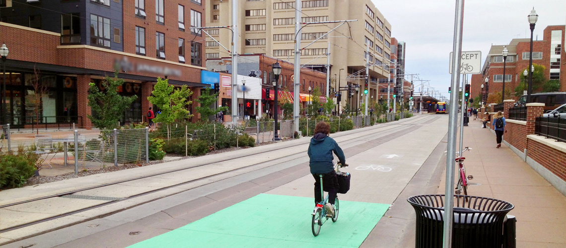 Photograph of a cyclist riding in a bicycle lane next to a row of businesses. A bus is visible in the background. 