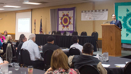 Photograph of HUD Secretary Julián Castro speaking at a podium to a roomful of people seated around tables.