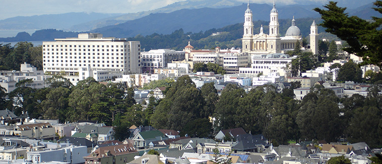 View of San Francisco showing homes and vegetation surrounding large, multistory buildings. Mountains are visible in the background. 