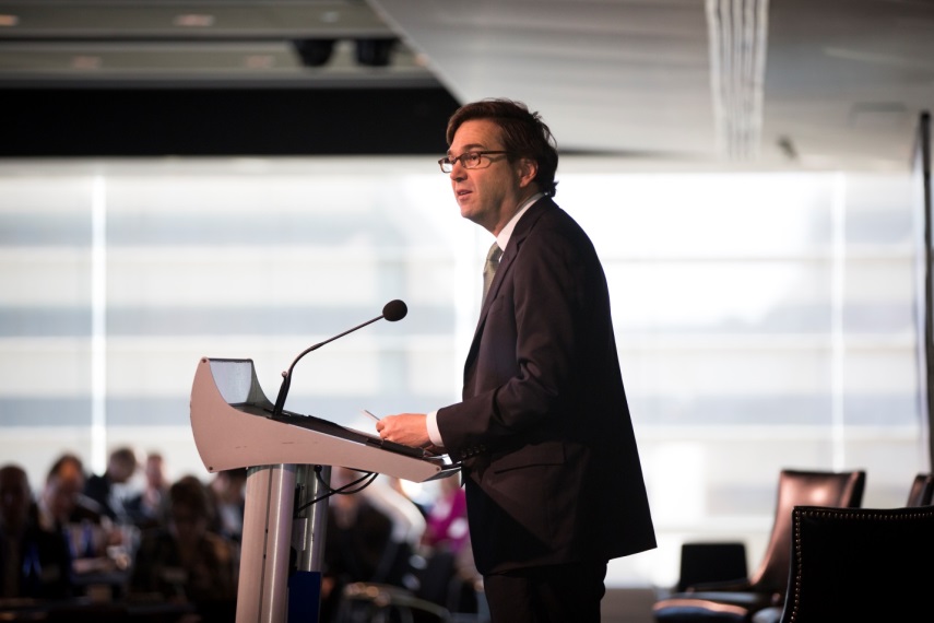 Photograph of Jason Furman speaking at a podium on a stage. A crowd people is visible in the background.