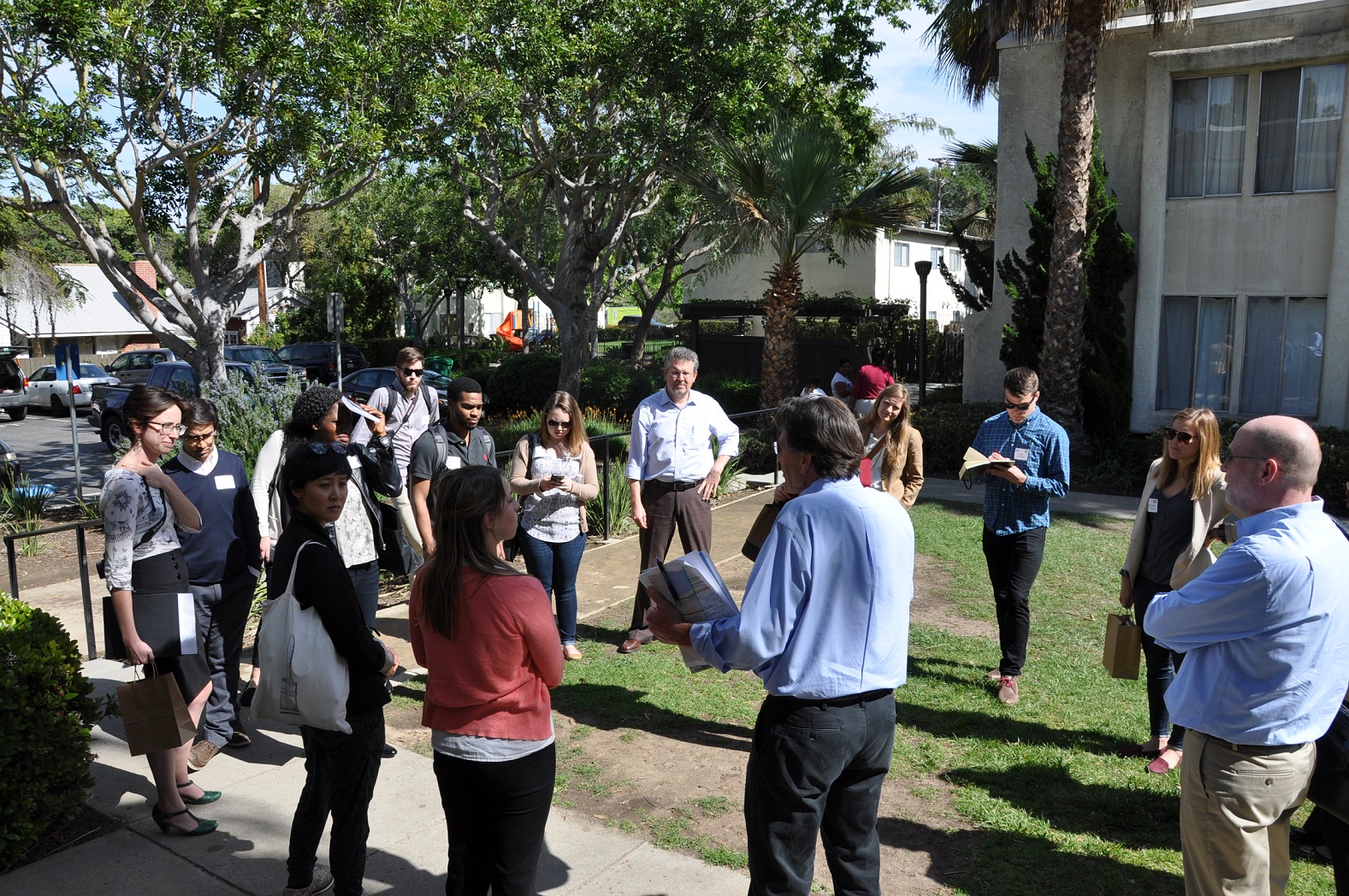 The finalist teams gather in a circle in a landscaped area near several multistory buildings.