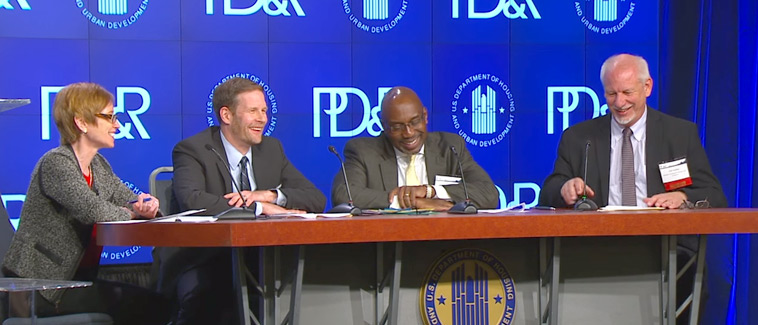 Moderator Katherine O’Regan and three panel participants sit behind a table in front of a background with the PD&R logo.