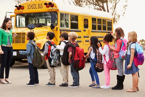 Young children wait in line in a parking lot with a school bus in the background.
