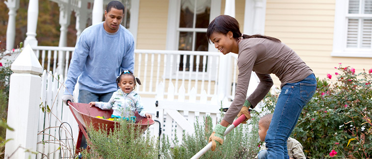 Image of a family landscaping the yard in front of a home. A woman using a gardening tool and a child sitting near her are shown in the foreground, and a man, pushing another child in a wheelbarrow, is visible in the background.