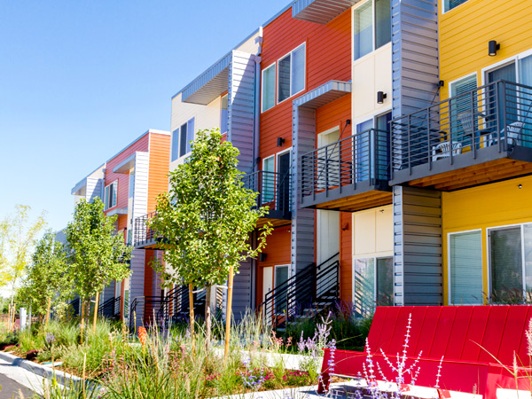 A bench and trees line the street in front of contemporary, two-story, multifamily housing units.