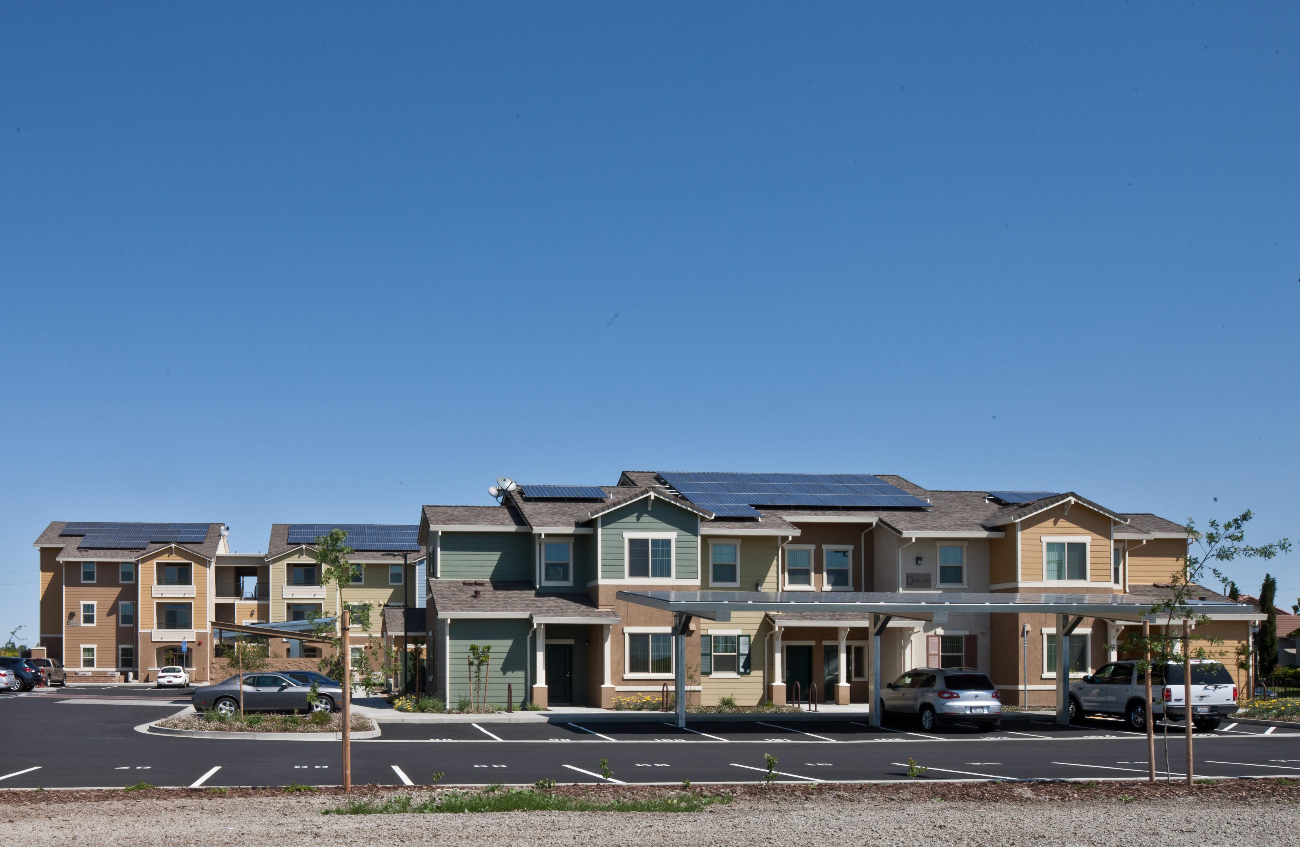 Photograph of the front façades of a two-story and a three-story apartment building with photovoltaic panels on the roofs. Panels are also installed over some of the parking spaces near the buildings.