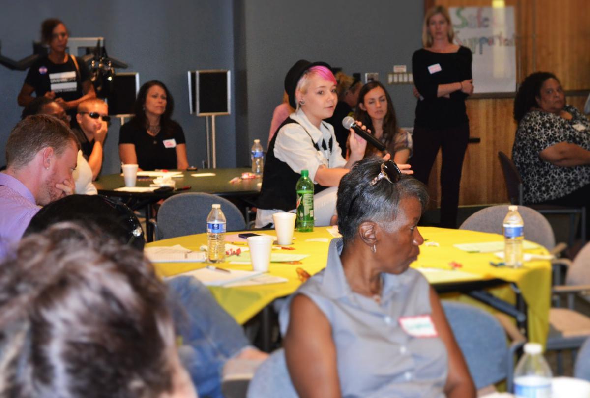 Photograph of a young adult speaking into a microphone and approximately a dozen other people sitting around several tables or standing in the back of a large meeting room. 