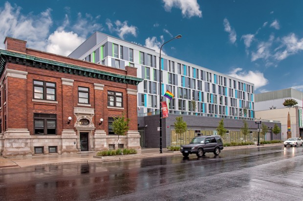 Photograph of the front of two buildings, a two-story brick structure and a new six-story steel and concrete apartment building next door. A rainbow flag is attached to a street light pole in front of the brick building, and the flag colors are incorporated into a sculpture at the far end of the new building.