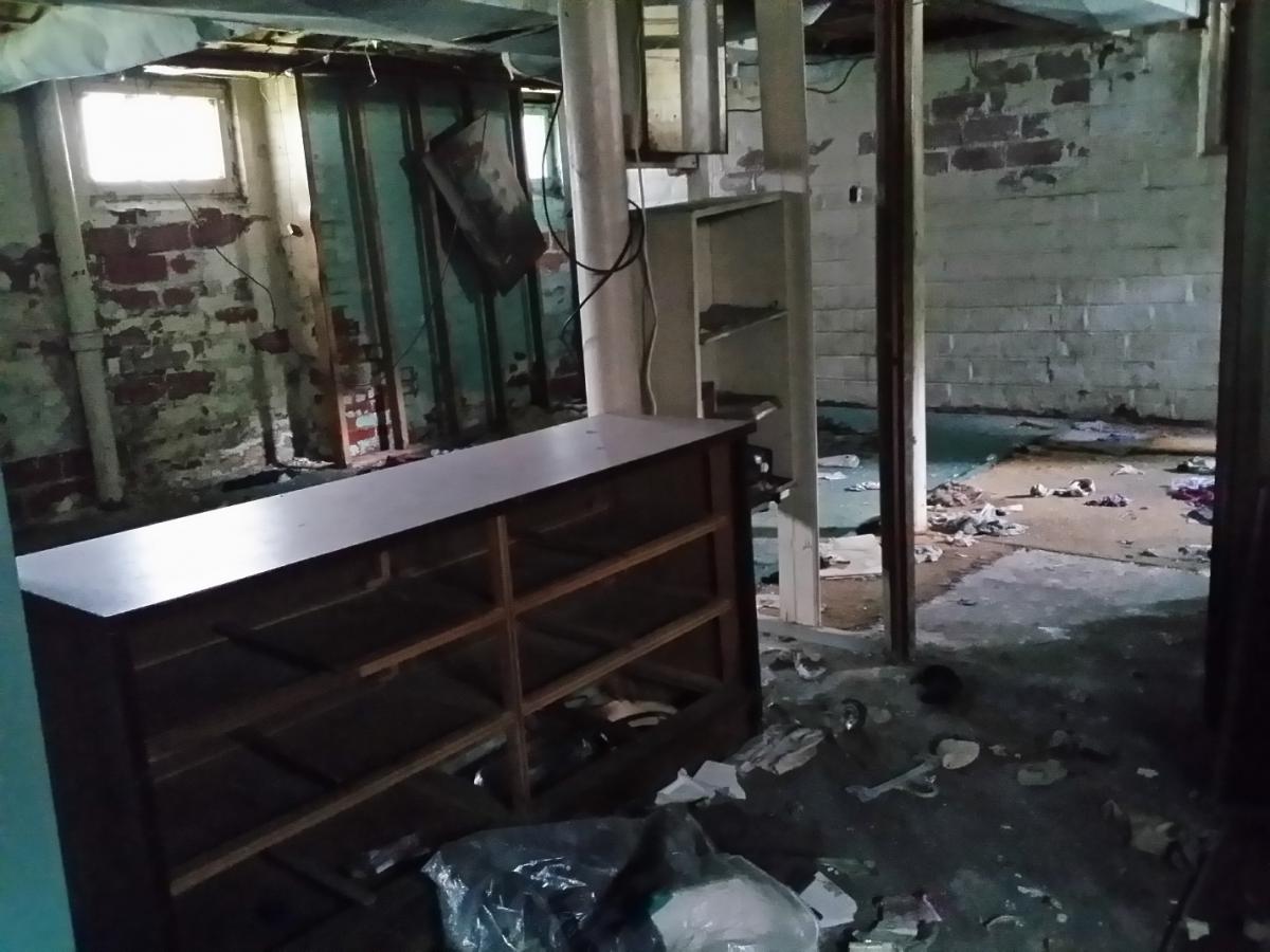 Photograph of a basement in a house prior to renovation with exposed studs and wires, along with damaged furniture and trash.