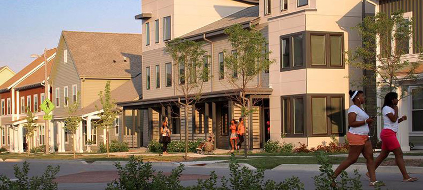 Several low-rise, multi-story buildings along a road, with individuals visible on a porch and walking along the road. 