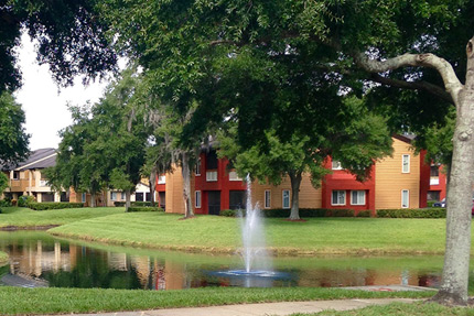 Photograph of multiple low-rise apartment buildings surrounded by landscaped grounds. A pond with a fountain is visible in the foreground. 