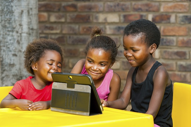 Photograph of three children, two boys and one girl, sitting at a table while viewing an electronic tablet.