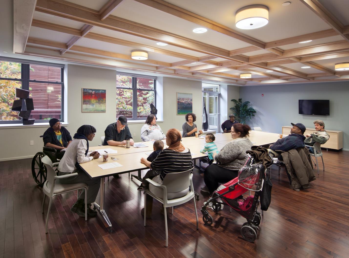 Photograph of ten adults and two toddlers sitting and having a discussion around a table in a modern conference room.