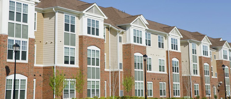 Photograph of a low-rise, multifamily residential building with a façade that includes siding and brick. Tress lines the front of the building.