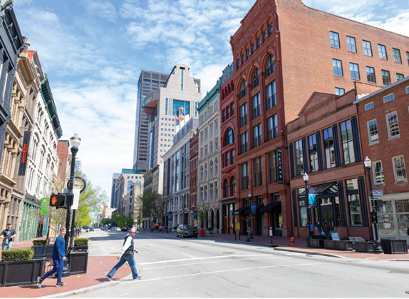 Photos shows buildings on either side of a street in downtown Louisville, Kentucky, with two pedestrians crossing a street in the foreground.