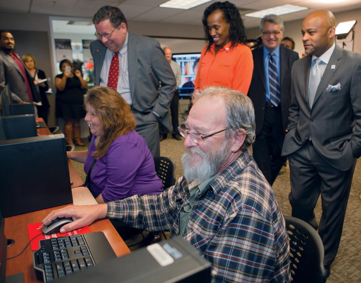 A man and a woman are seated in front of computers with four other adults standing behind them.