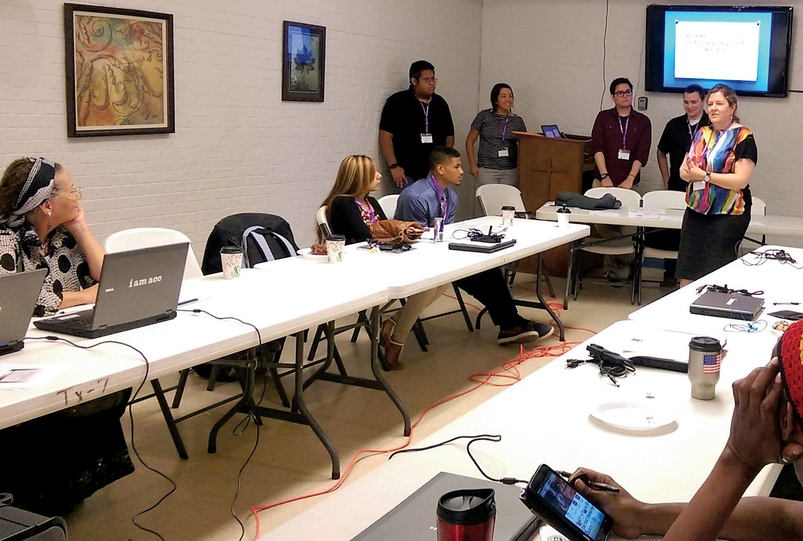 A group of people, some seated at long narrow tables with laptop computers, listen to a presentation being given by a woman.