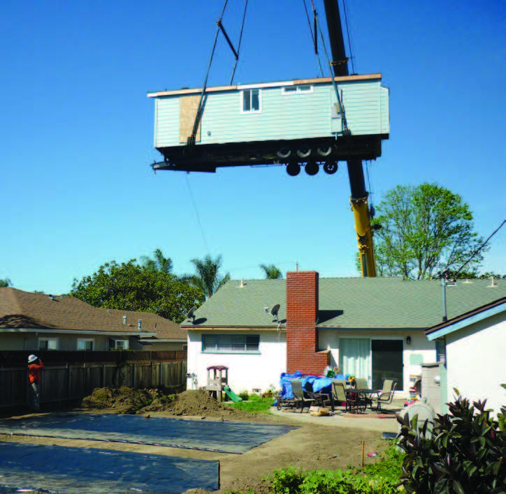 Photo shows a factory-built manufactured home held by a crane in the air above the backyard of a one-story, single-family home.