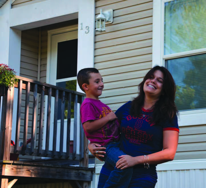 Smiling woman standing holding a child with the front wall and door of a home behind her.
