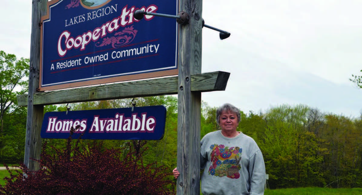 A woman standing next to a wooden sign with the text “Lakes Region Cooperative” and “Homes Available” with trees in the background.