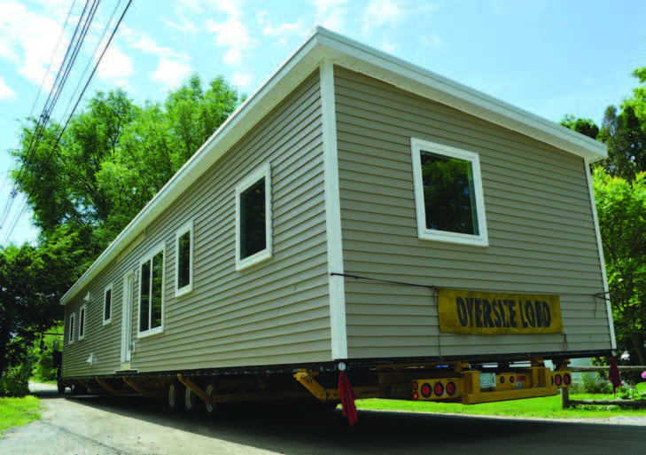 Rear and side view of a modular home being transported on a flat bed truck on a neighborhood street.
