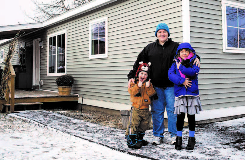 A woman and two children stand in front of modular home with snow on the ground.