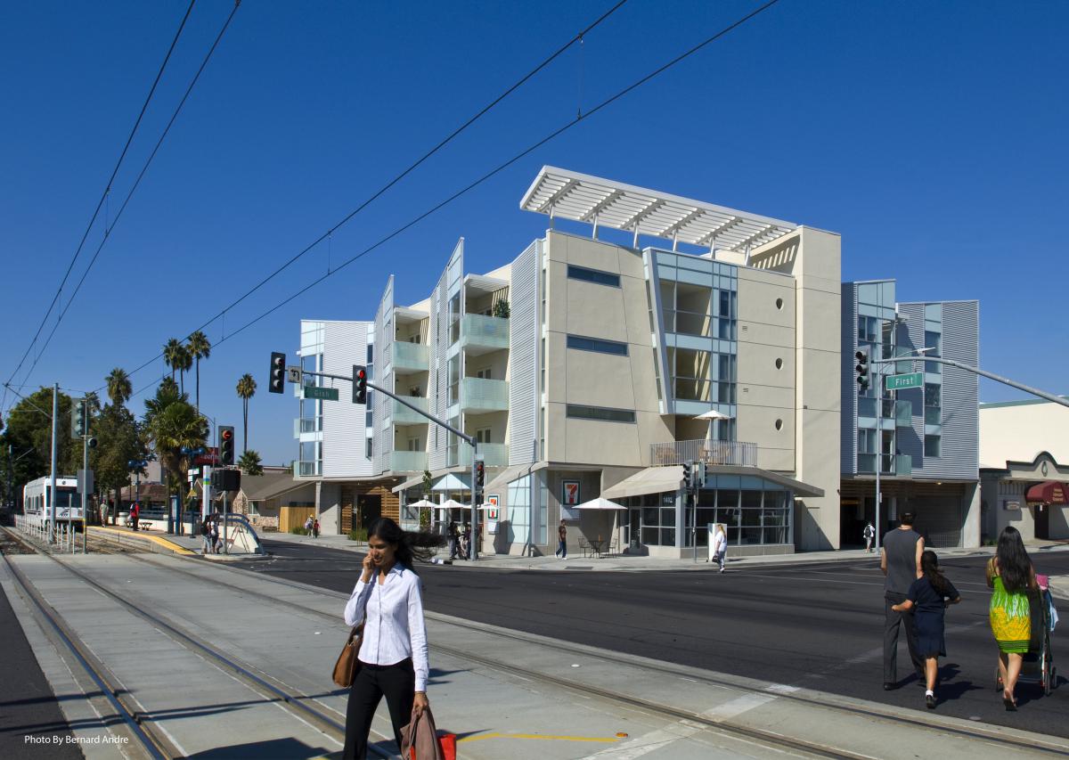 A four-story apartment building with pedestrians walking across rail tracks located in front of the building.