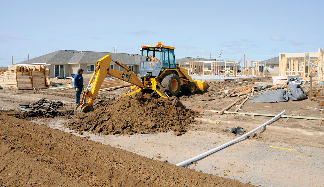 Photo shows two men, one operating a backhoe, with homes under construction in the background.