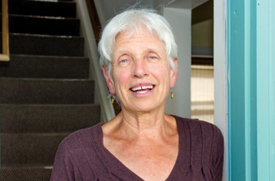 An elderly woman stands in the doorway of her home with a flight of stairs in the background.