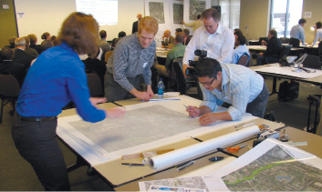 Photo shows four adults standing around a table with a large map on it with another larger group of adults watching a presentation on a screen in the background.