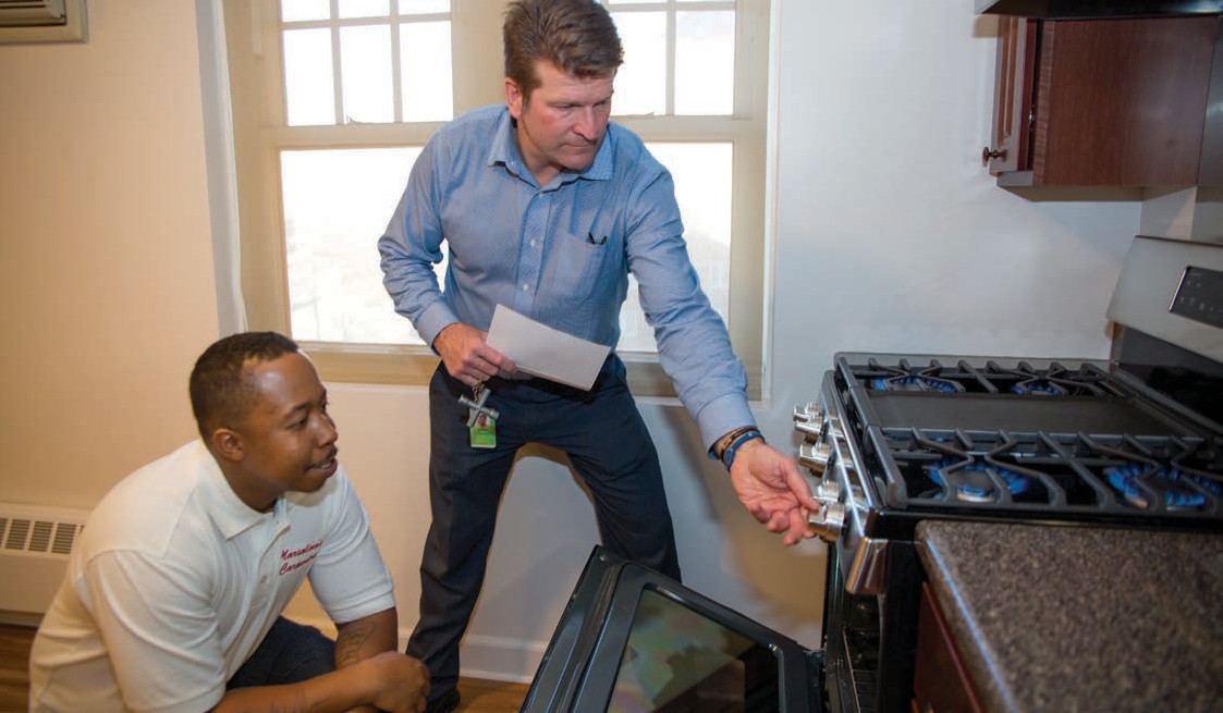 Man kneeling in front of a range stove watching another man demonstrating operation of the stove.