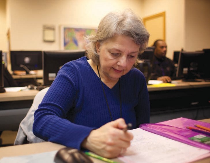 A woman seated at a table writing in a folder with rows of desktop computers in the background.