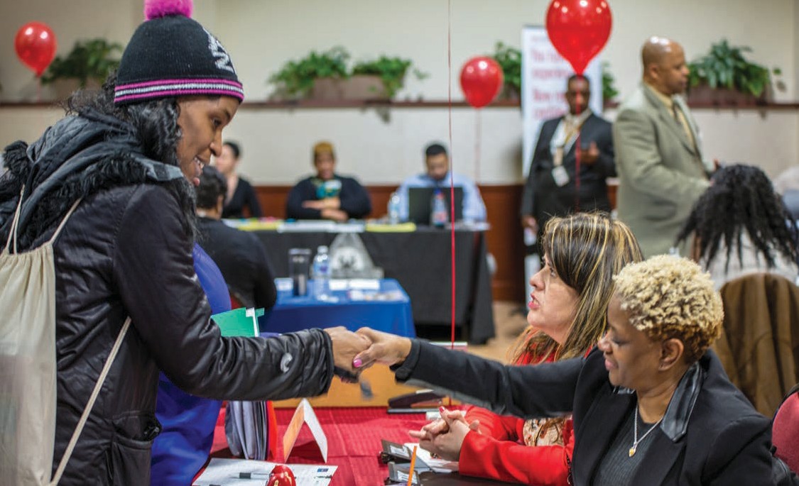 A woman stands in front of a table shaking hands with two women seated on the other side of the table.