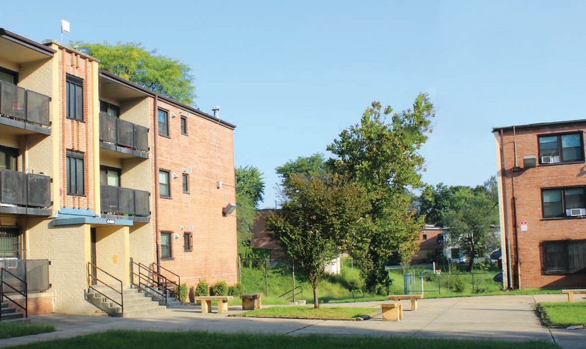 Two, 3-story apartment buildings situated perpendicular to each other with open space in between and a paved area with benches in the front.