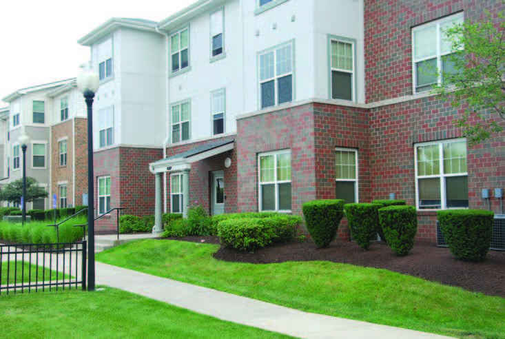 A three-story multifamily building with sidewalk and landscaping in the foreground.