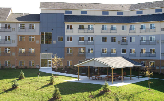 A five-story building with landscaping and a covered outdoor area with benches in the foreground.