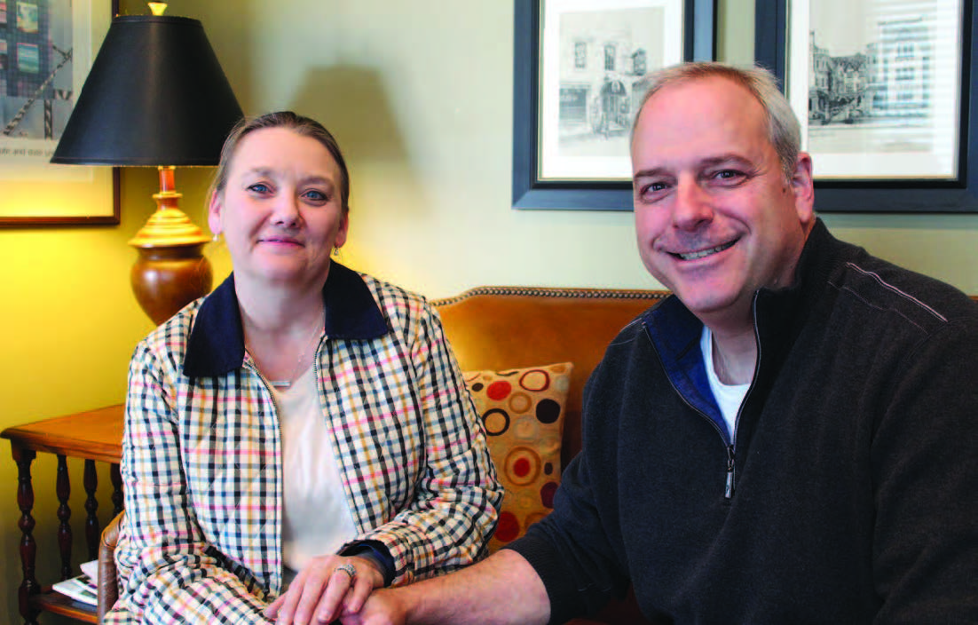 Photo of a man and a woman seated on a sofa holding hands and smiling into the camera.