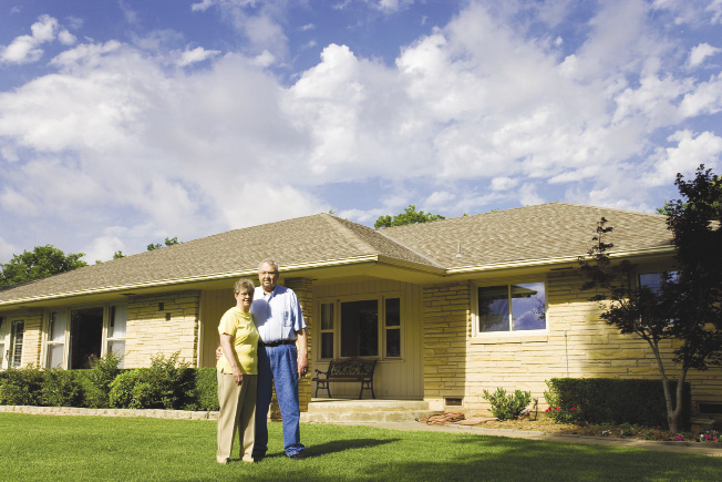 Photo shows an elderly couple standing in front of their home.