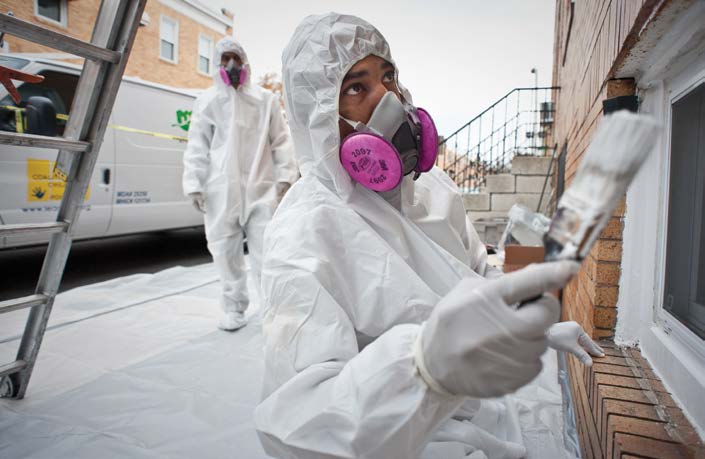 A man wearing protective gear is standing on a sidewalk painting a window frame.