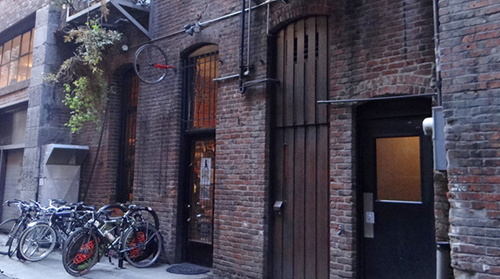 Photograph of an alley in which several bikes are parked adjacent to a brick building with multiple doors.