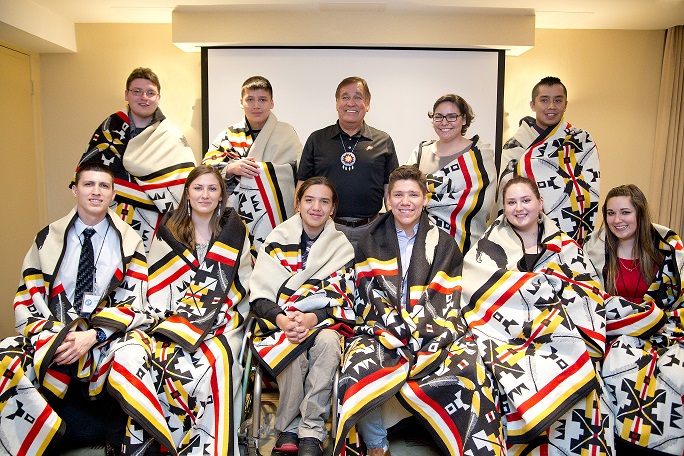 Image of the 10 young adults participating in Dreamstarters, wrapped in colorful blankets, sitting or standing near the program’s creator, Billy Mills.