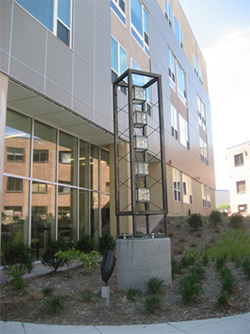 Photograph of a sculpture located in a garden near the entrance of a four-story multifamily apartment building.