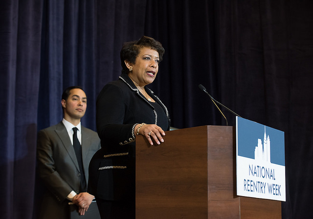Photograph of Attorney General Loretta Lynch speaking at a podium bearing the National Reentry Week logo. HUD Secretary Julián Castro is visible onstage in the background. 
