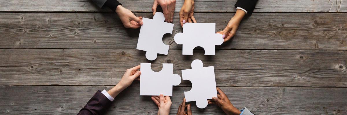 Four people holding large white puzzle pieces to represent cooperation and unity.