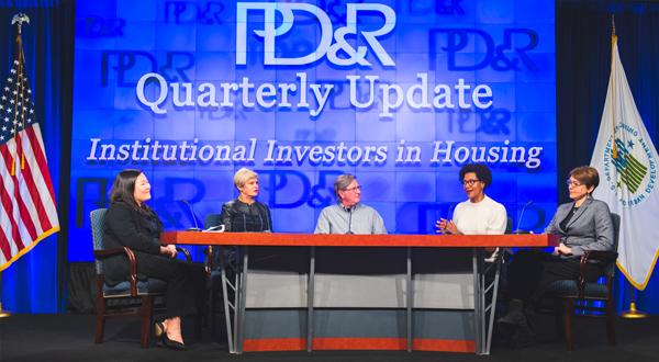 Researchers and community practitioners discussing the Quarterly Update around a table. A blue screen behind them has the text 'PD&R Quarterly Update Institutional Investors in Housing'.