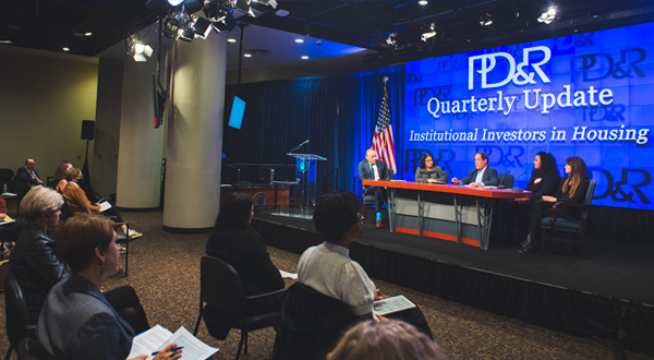 A group of people sit and listen to speakers sitting around a table. A blue screen behind the speakers has text that reads 'PD&R Quarterly Update Institutional Investors in Housing'.